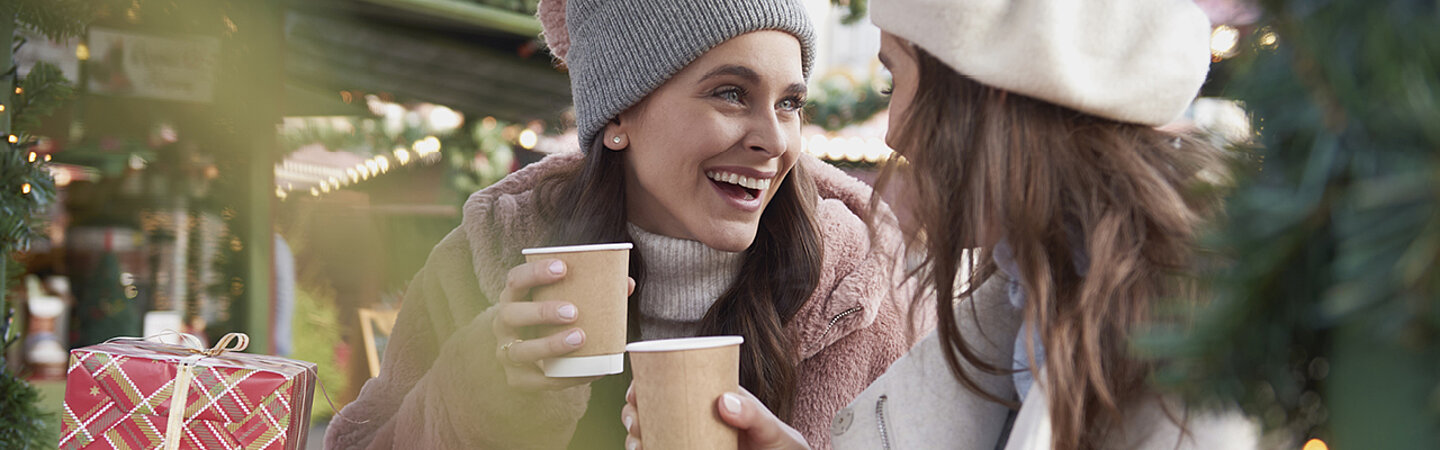 Zwei Frauen an einem Weihnachtsmarkt trinken Heissgetränke aus Getränkebechern aus nachhaltigem Kraft-Karton