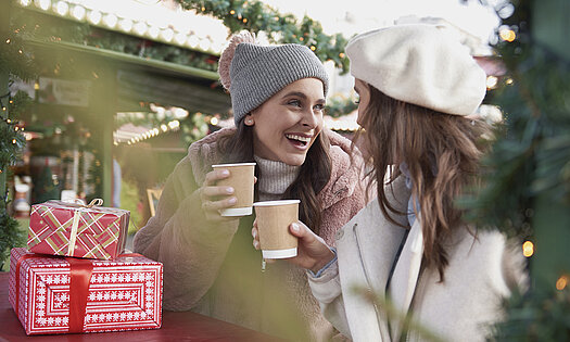 Zwei Frauen an einem Weihnachtsmarkt trinken Heissgetränke aus Getränkebechern aus nachhaltigem Kraft-Karton